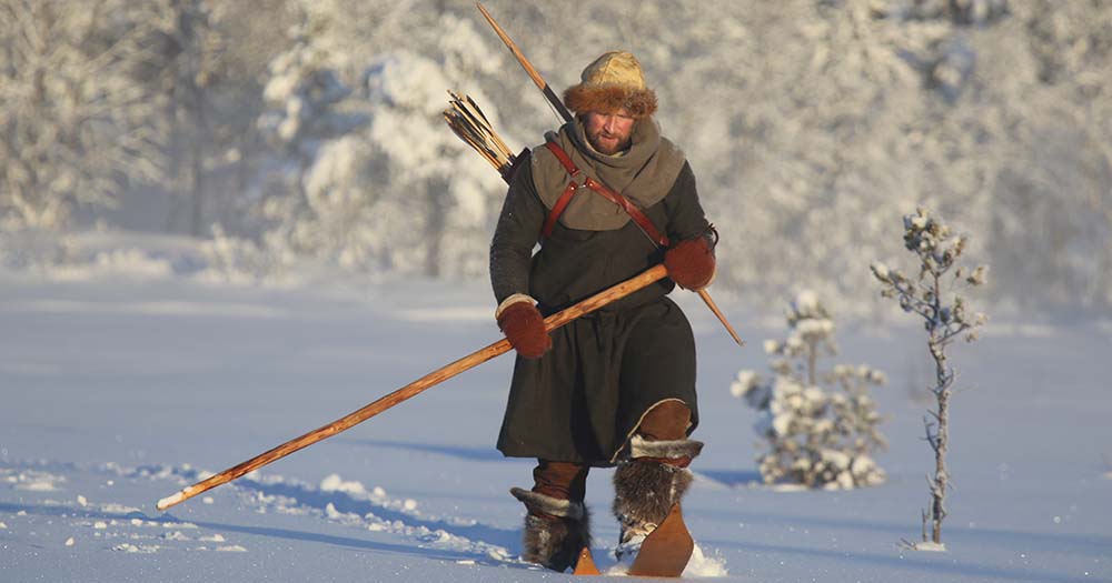 Mann ikledd gammeldagse skinnklær går i snøen med jegerutstyr.