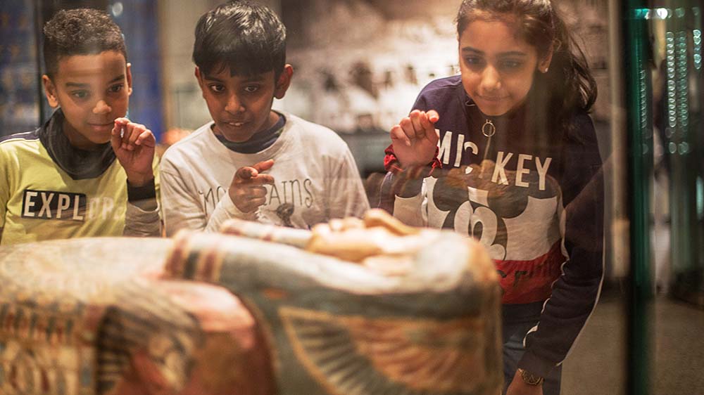 To gutter og en jente studerer en sarkofag gjennom en monter.
