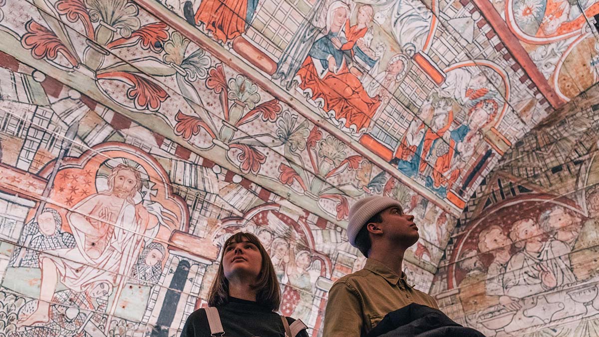 Two people looking up at colorful paintings on a large wooden roof 
