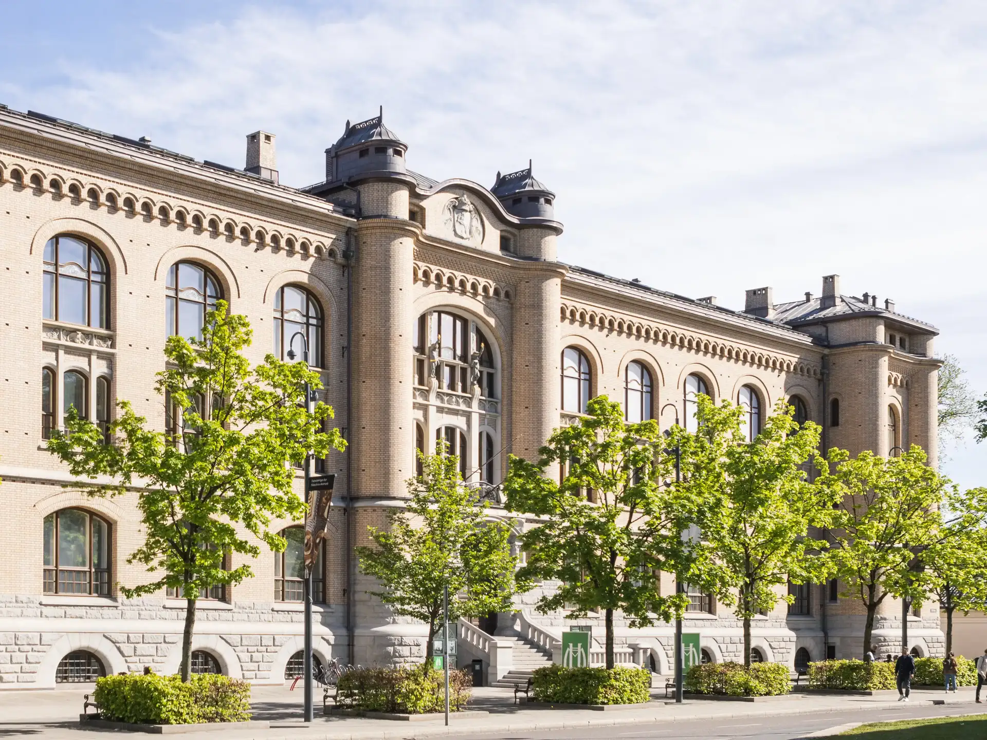 Foto av fasaden på et stort, flott museumsbygg i stein. Blå himmel med skyer, en rekke med grønne trær står foran bygget