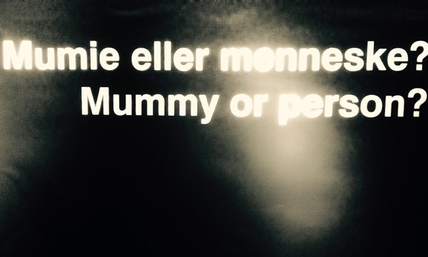 Svart bakgrunnsfarge, hvit tekst "Mumie eller menneske? Mummy or person?"