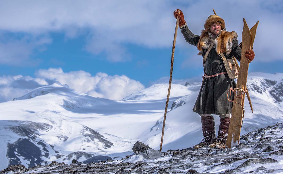 Man in Viking clothes standing on top of a snowy mountain looking at the majestic view. Holding skis and ski poles